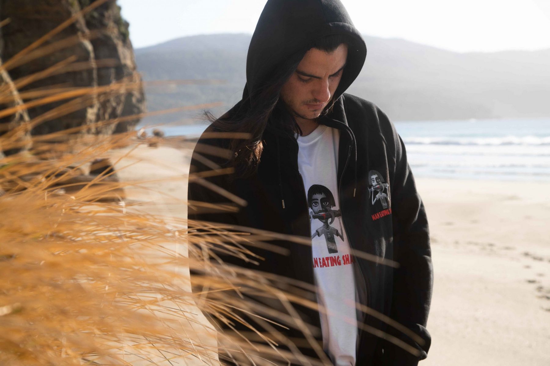 Man wearing Man Eating Shark t-shirt in white with black hoodie standing on beach Tasmania