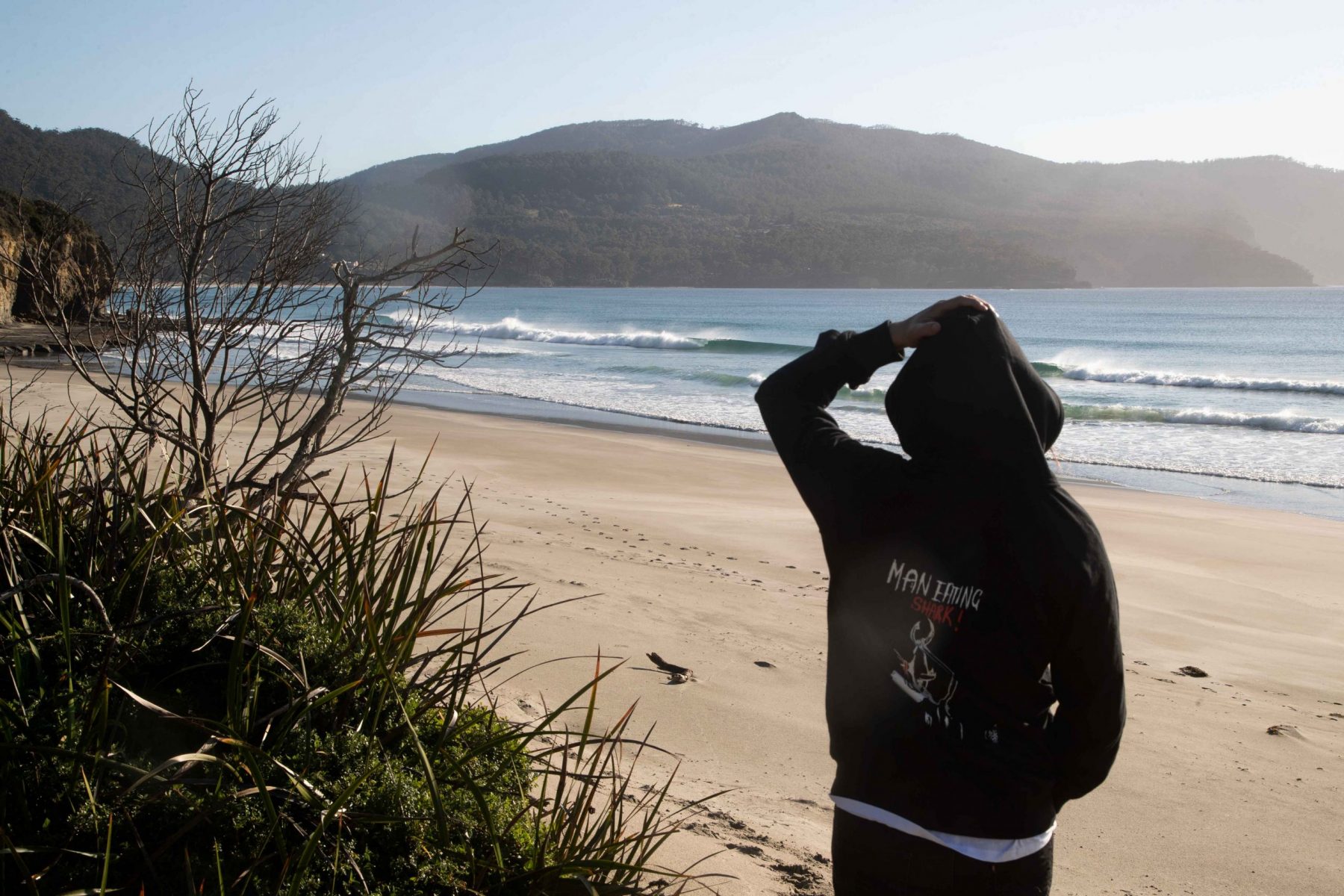 Man with his hand on his head wearing black Man Eating Shark Hoodie with Punk design beach in the background