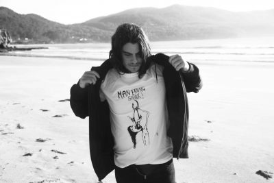 Man putting on black hoodie over white long sleeved Man Eating Shark Punk t-shirt with Tasmanian beach in the background